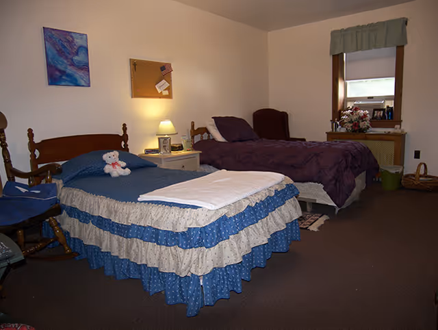 A cozy bedroom with two single beds, one with a blue and white ruffled bedspread and a teddy bear on it, the other with a dark purple quilt. Between the beds is a nightstand with a lamp, picture frame, and some small items. There is a wooden rocking chair to the left, a window with a green valance on the right wall, and a small table with flowers and other items beneath the window. The walls are light-colored and the floor is carpeted.