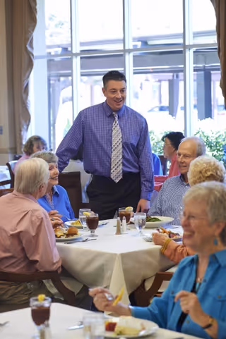 A group of elderly people sitting around a dining table in a well-lit room, enjoying a meal and drinks. A man in a purple checkered shirt and patterned tie is standing and engaging with the group, smiling. Large windows in the background let in natural light.