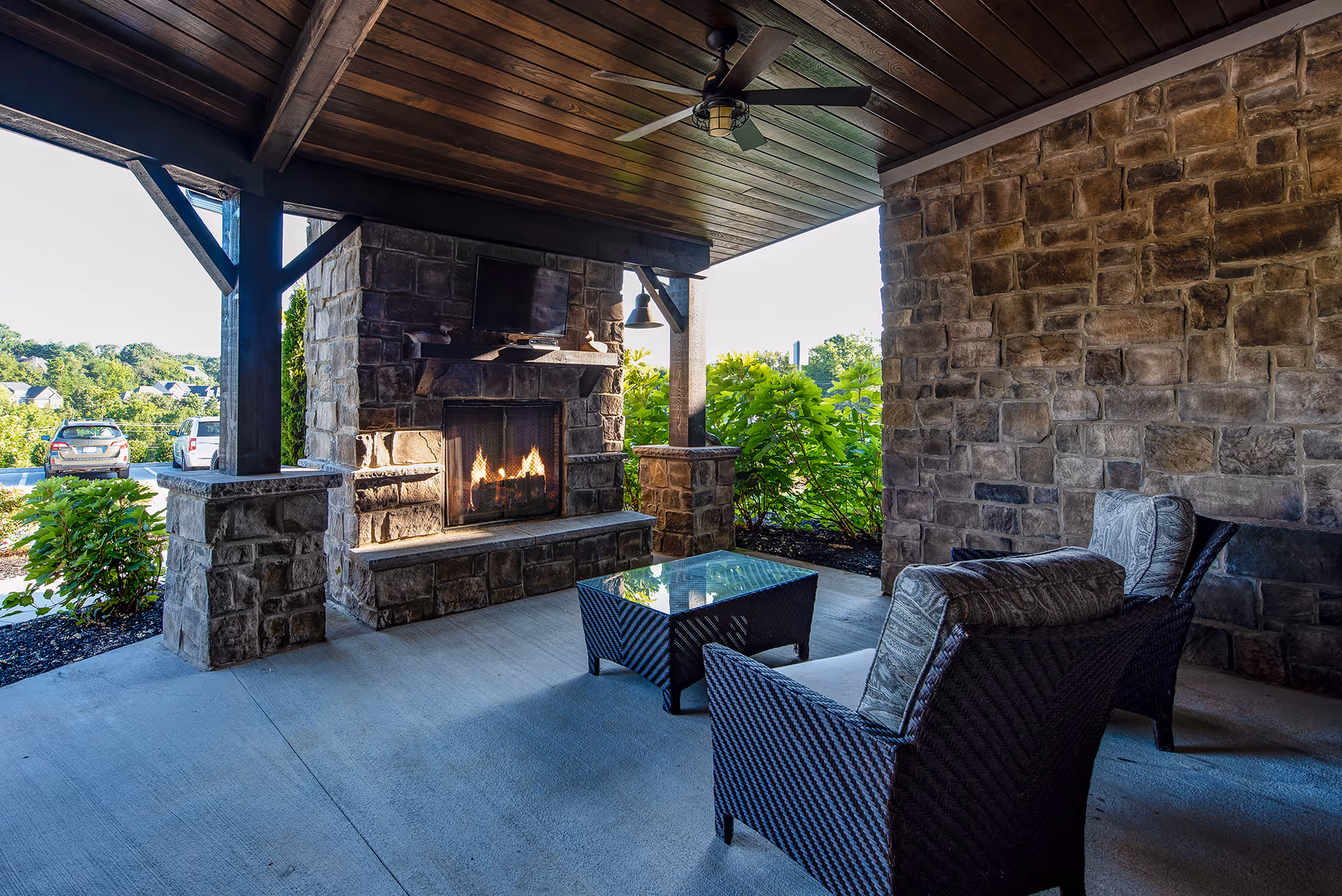 Covered outdoor seating area with a stone fireplace, wicker chairs, and a glass-top coffee table overlooking greenery.