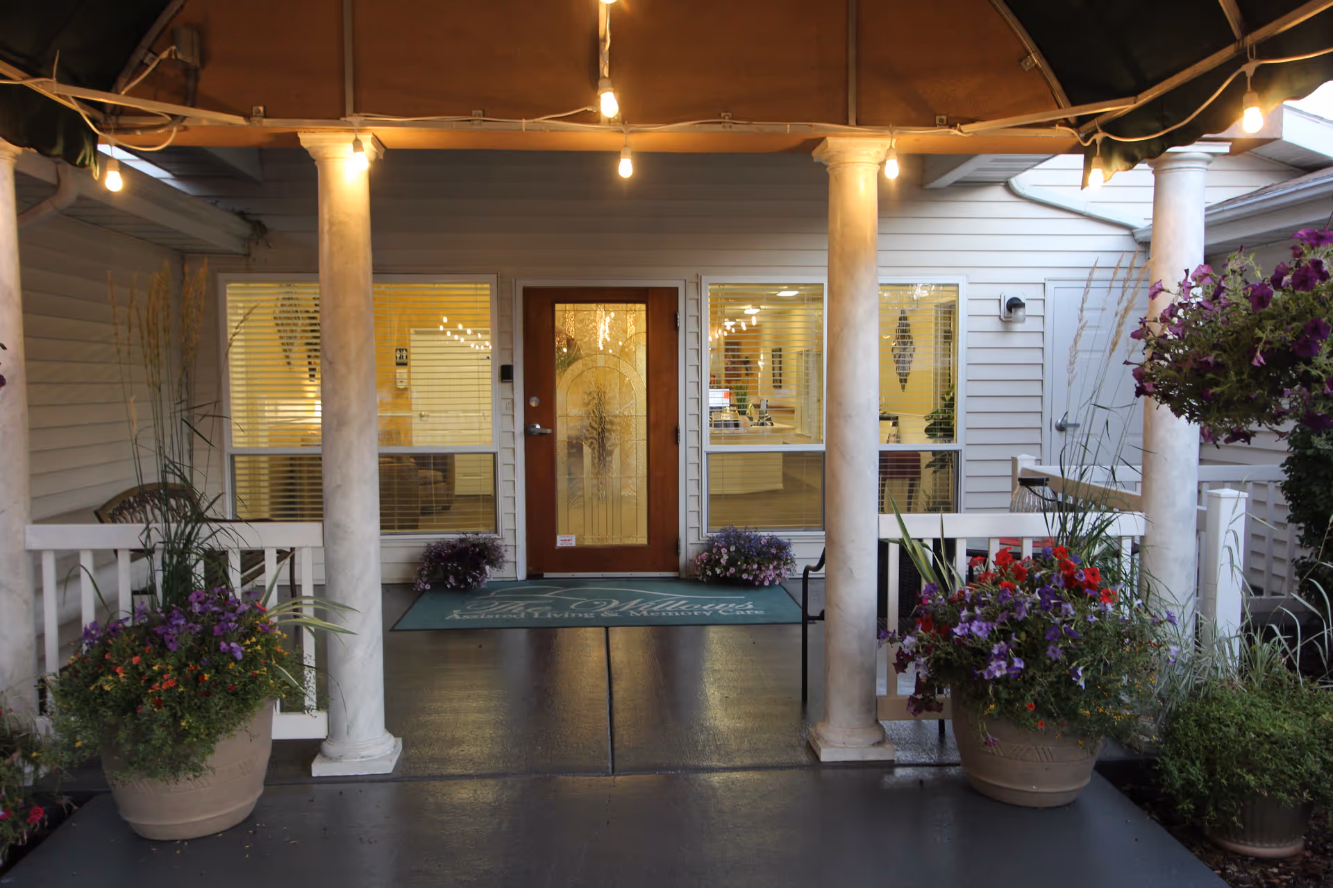 Entrance to The Willows Retirement & Assisted Living facility with a covered porch supported by white columns, potted plants with flowers on either side, and a glass door with decorative design. The interior reception area is visible through the windows.