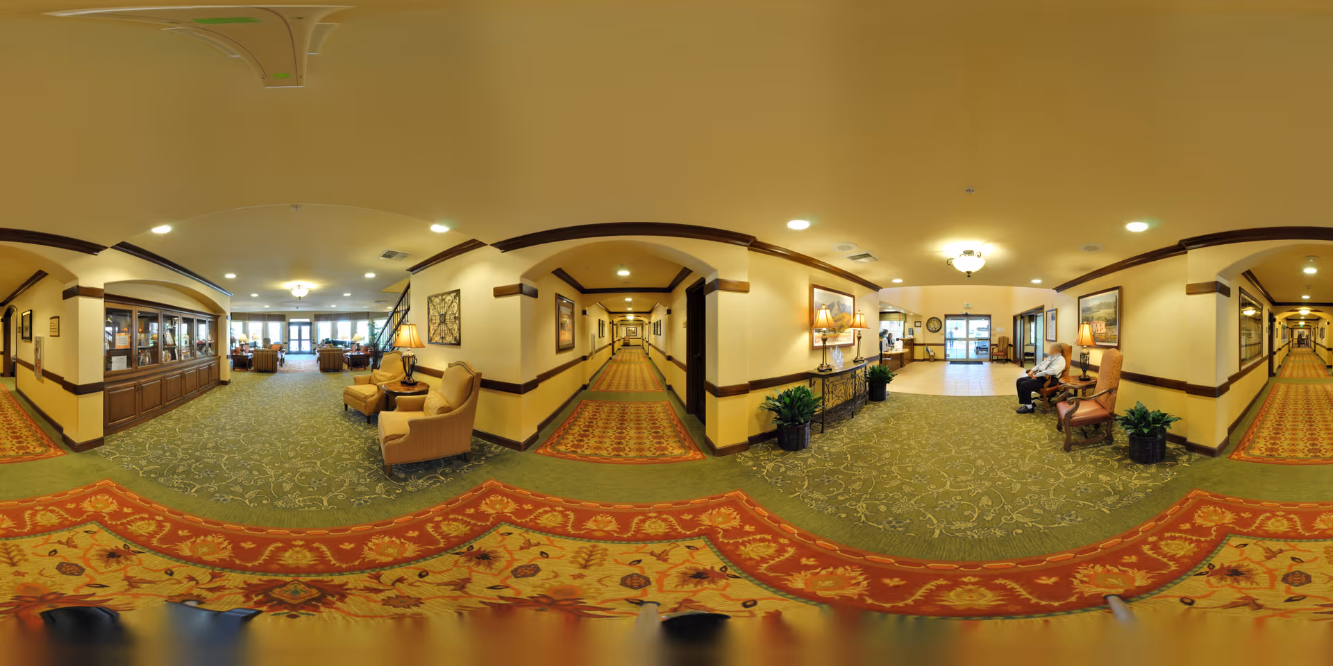 Panoramic interior of a senior living facility lobby with seating areas, patterned carpets, plants, and long hallways.