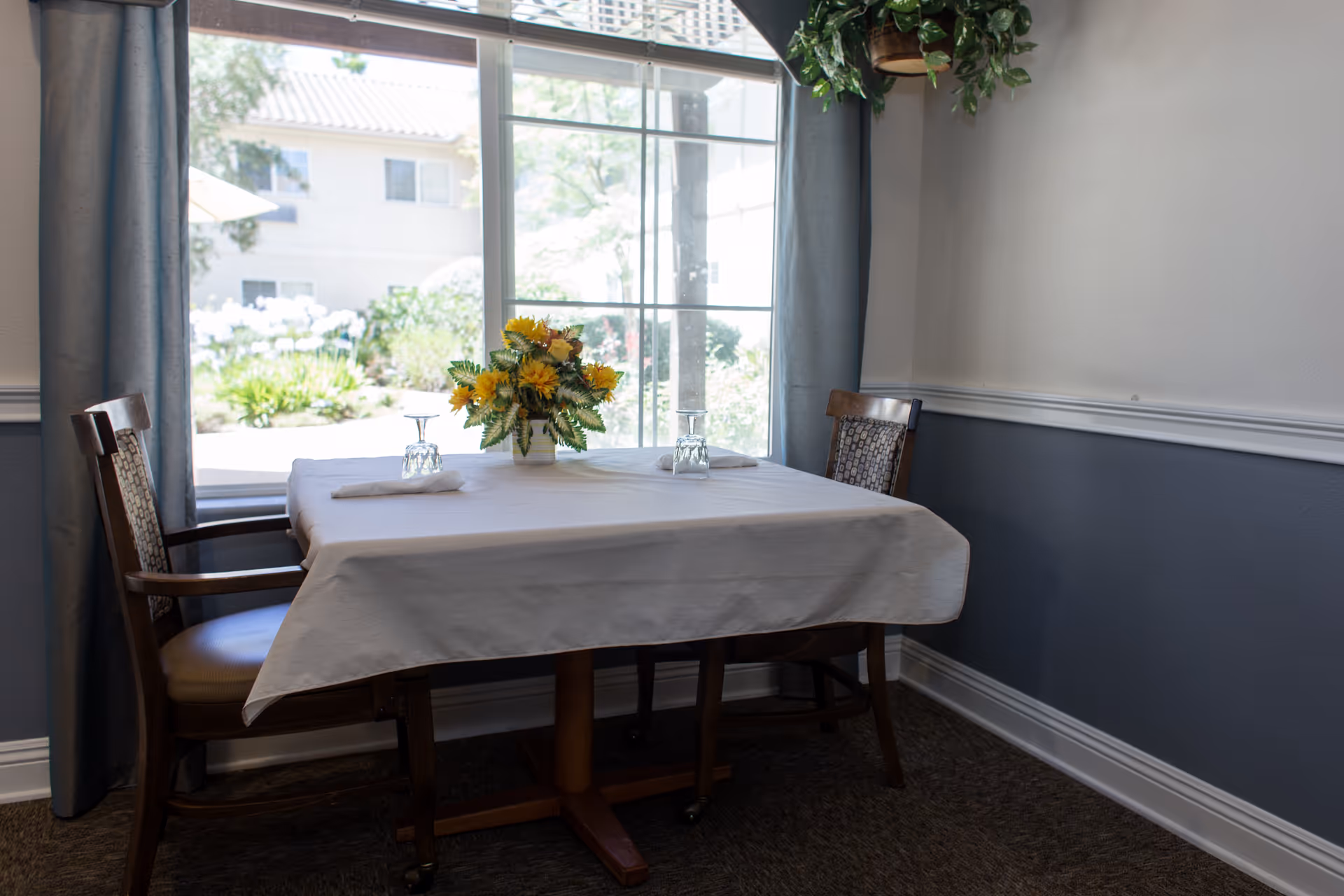Small dining table set for two with a white tablecloth, a floral centerpiece and two chairs in front of a large window.