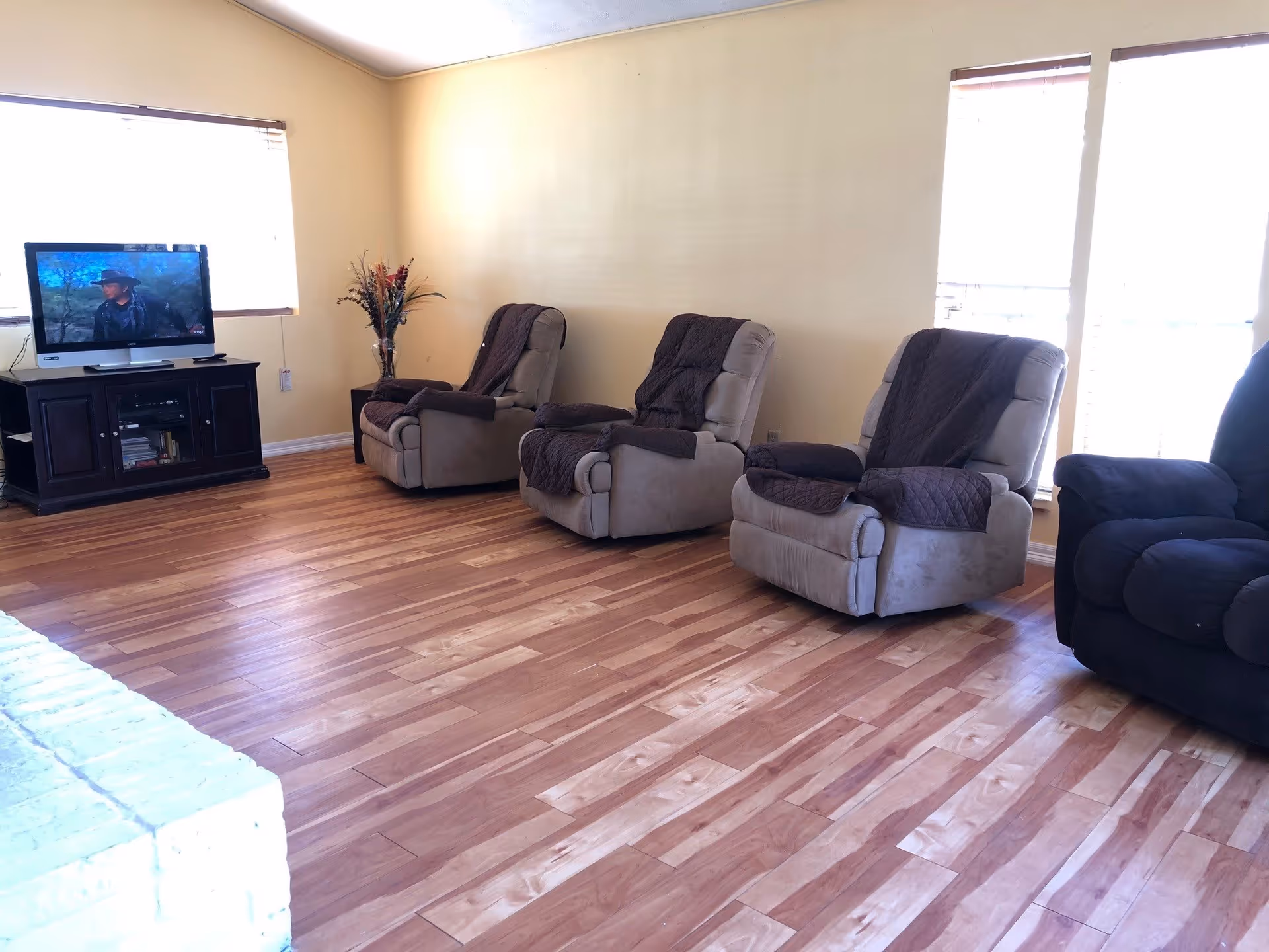 A living room with four recliner chairs, three beige and one dark blue, each with a dark brown throw blanket draped over the back. There is a wooden TV stand with a flat-screen TV showing a scene with a man wearing a hat. The room has light yellow walls, two windows with blinds, and a wooden floor.