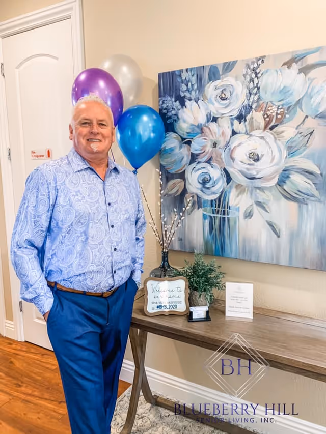 An older man wearing a light blue patterned shirt and blue pants stands smiling next to a wooden console table with decorative items including a vase with branches, a small plant, and a sign. Behind the table is a large painting of white and blue flowers. There are also purple, silver, and blue balloons attached to the table. The setting appears to be indoors with a wooden floor and a closed white door in the background.