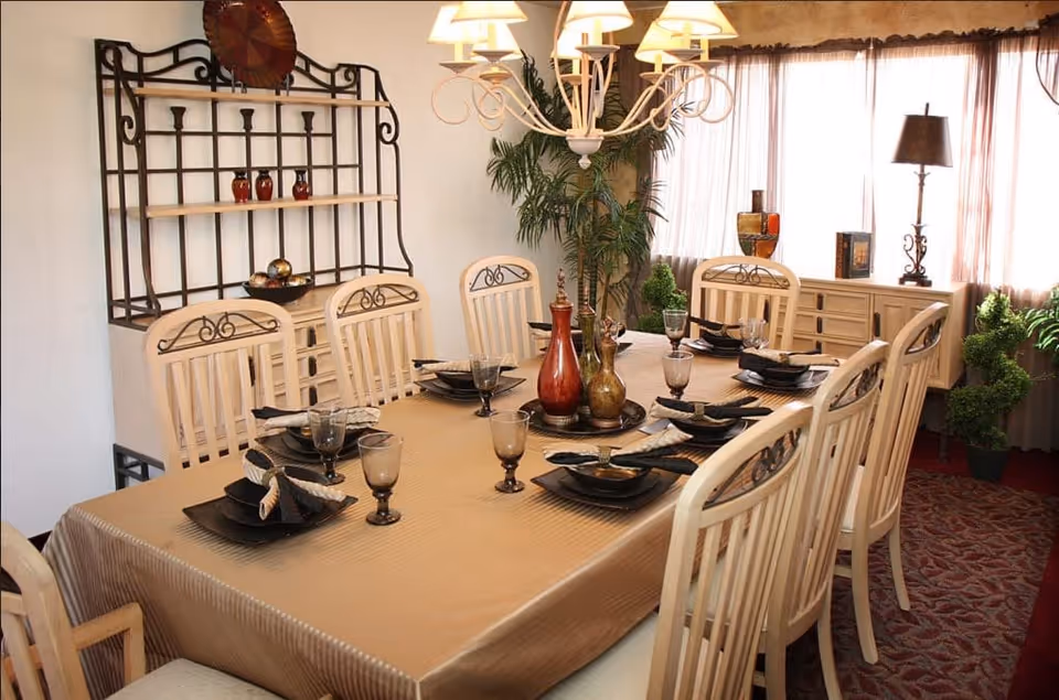 A dining room with a long rectangular table covered with a beige tablecloth, set with black plates, glasses, and folded napkins. Eight wooden chairs with decorative backs surround the table. A chandelier with multiple lampshades hangs above the table. In the background, there is a decorative metal shelf with small vases, a large potted plant, a sideboard with decorative items, and a floor lamp near a window with sheer curtains.
