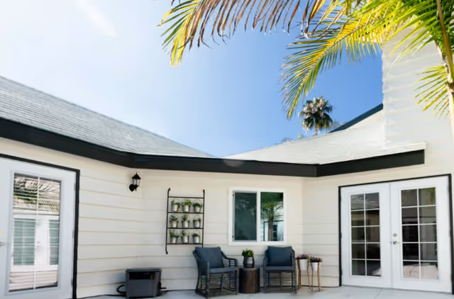 Bright exterior courtyard with white siding, two wicker chairs and a small table beneath palm fronds in front of French doors.