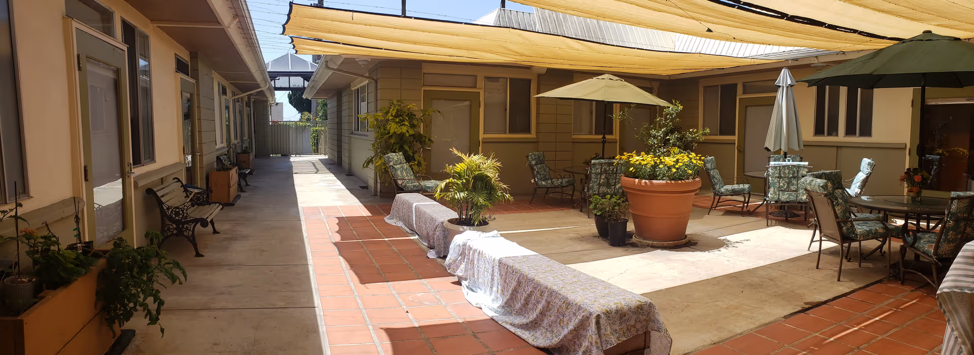 Outdoor courtyard area at Ventura Villa Assisted Living with benches covered in fabric, potted plants, patio tables and chairs with umbrellas, surrounded by single-story building walls and windows.