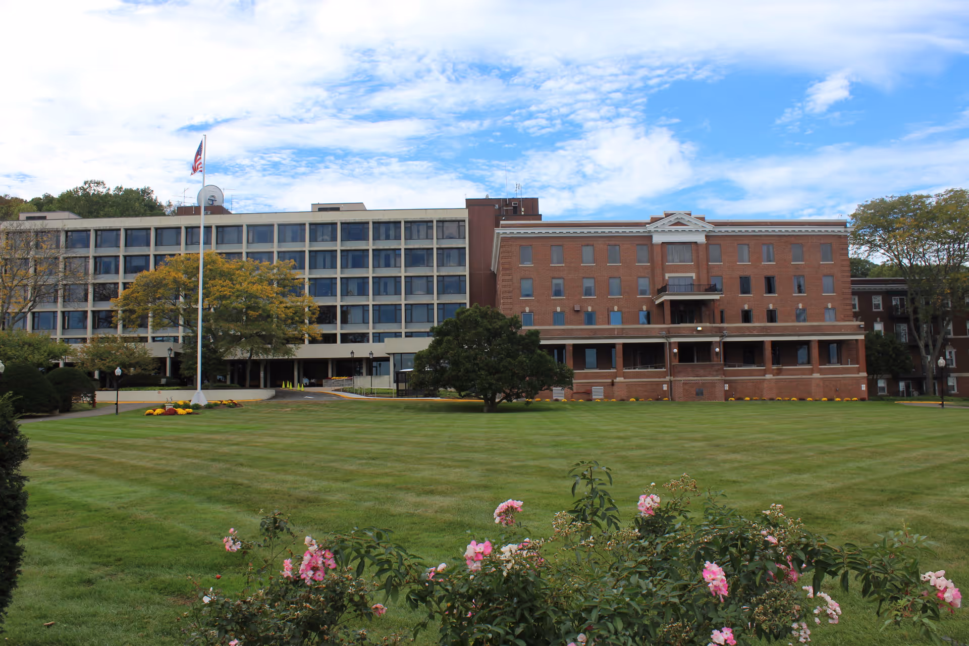 Exterior view of Masonicare Health Center showing a large multi-story building with a combination of modern and traditional brick architecture. The building is surrounded by a well-maintained lawn with a flagpole flying the American flag and some flowering bushes in the foreground under a partly cloudy sky.