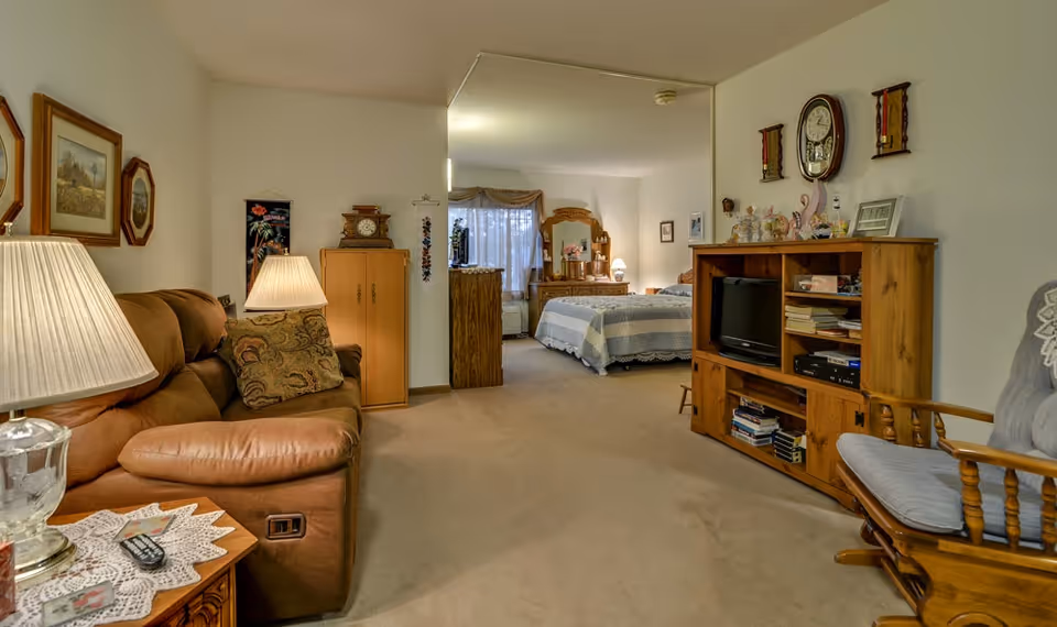 Interior view of a senior living facility room at River Bend showing a cozy living area with a brown recliner sofa, a wooden rocking chair, a wooden entertainment center with a TV and books, and a bedroom area in the background with a bed, dresser, and window with curtains.