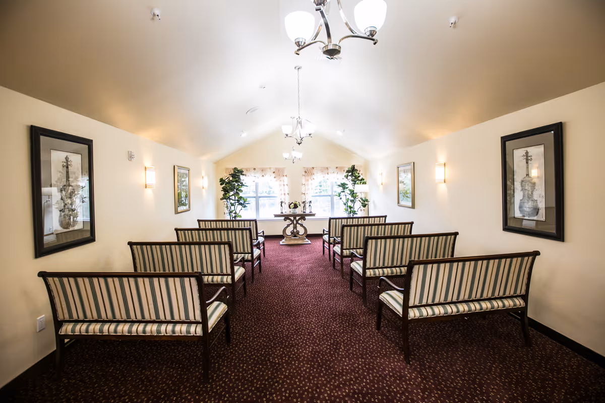 Interior view of a small chapel or meeting room with rows of wooden benches with striped cushions facing a table with decorative items in front of two large windows. The room has beige walls, framed artwork, wall sconces, and ceiling light fixtures.