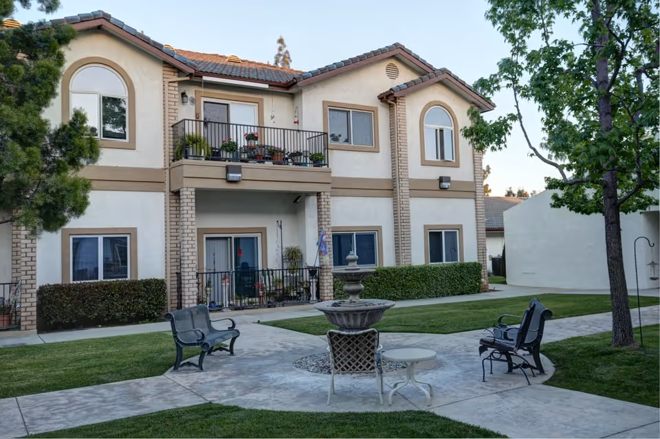 Outdoor courtyard area of a senior living facility with a two-story beige building in the background. The building has balconies with potted plants and several windows. In the courtyard, there are benches, a small round table with chairs, a decorative fountain, and trees providing shade. The area is surrounded by well-maintained grass and paved walkways.
