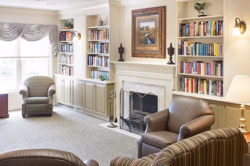 A cozy living room area with built-in bookshelves filled with books on either side of a white fireplace. There are several armchairs arranged around the fireplace, a large window with draped curtains letting in natural light, and a framed landscape painting above the fireplace.