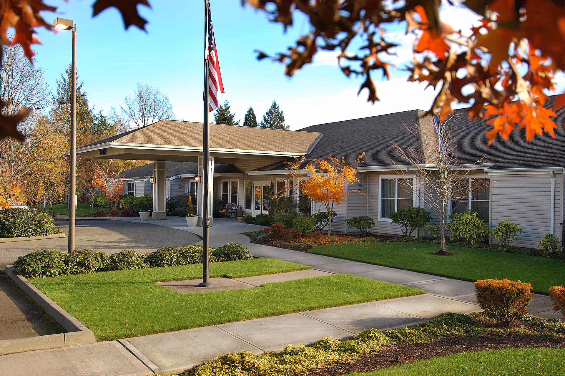 Exterior view of a single-story senior living facility building with a covered entrance, an American flag on a flagpole, well-maintained green lawns, shrubs, and trees with autumn foliage under a clear blue sky.