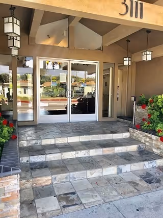 Entrance to a building with glass double doors, stone tiled steps leading up to the doors, hanging lantern-style lights, and potted plants with red flowers on either side of the steps.