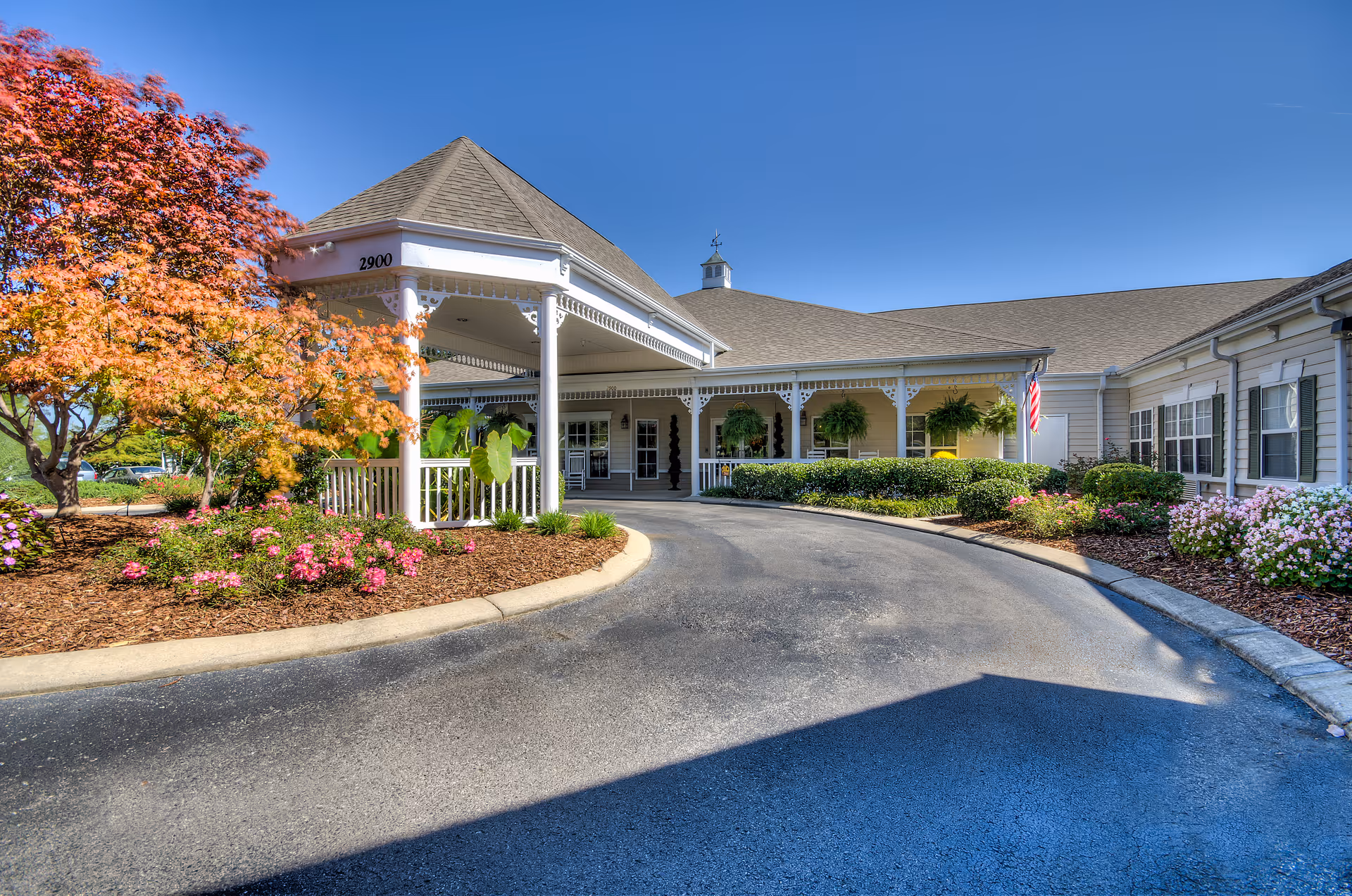 Exterior view of Charter Senior Living of Cleveland showing the entrance with a covered driveway, surrounded by well-maintained landscaping including flowering bushes and a tree with autumn-colored leaves under a clear blue sky.