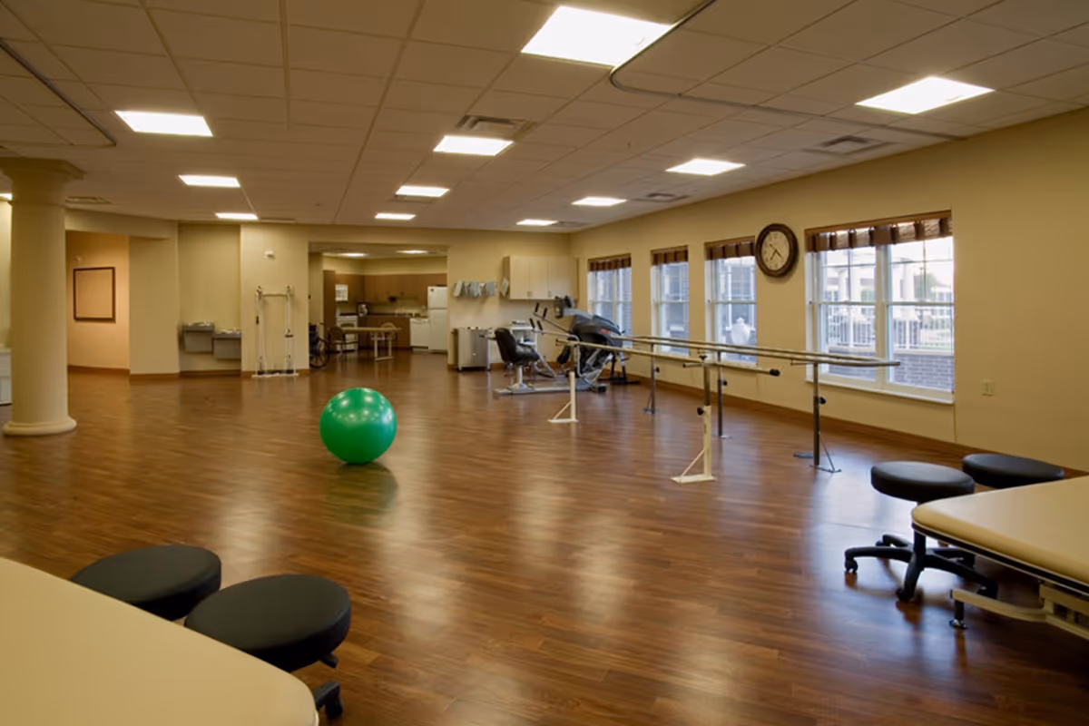 A spacious rehabilitation or physical therapy room with wooden floors, a large green exercise ball in the center, parallel bars for walking exercises along the right side near windows, exercise equipment, stools, and a clock on the wall. The room is well-lit with ceiling lights and has a kitchen area in the background.