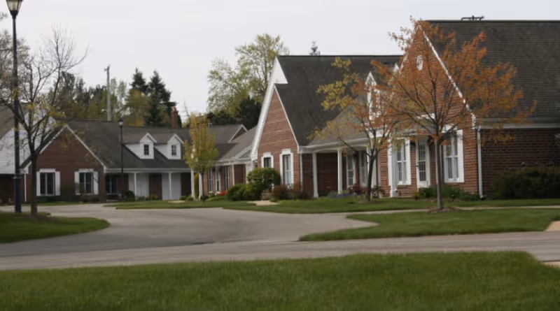 A quiet residential street in a senior living community with single-story brick houses, small trees with autumn leaves, and well-maintained lawns under an overcast sky.