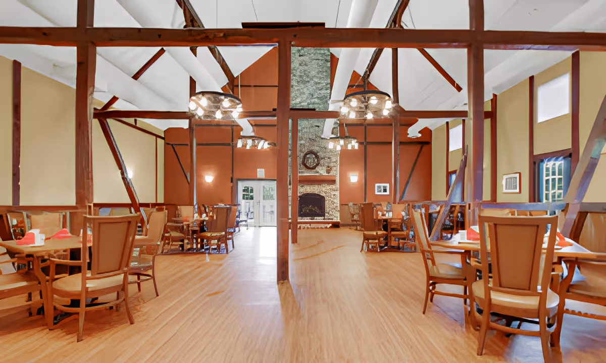 Spacious dining room with wooden floors, multiple tables and chairs, exposed wooden beams, and a large stone fireplace at the far end. The room has warm lighting and high ceilings with white panels.