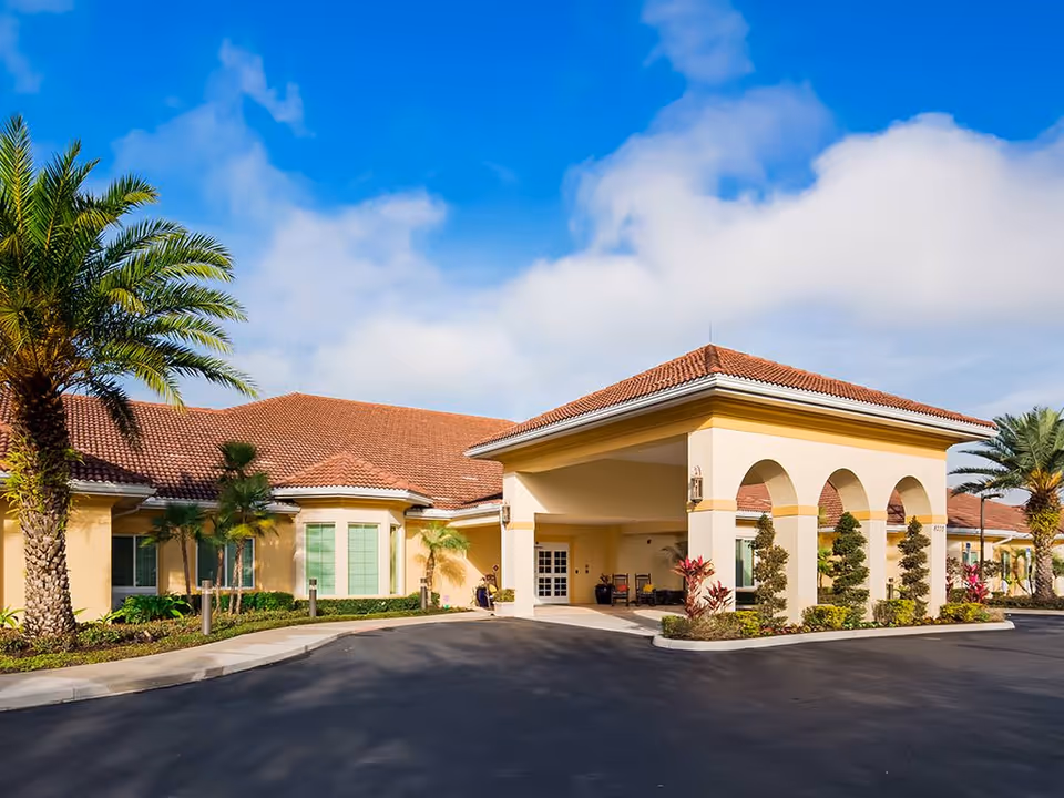 Mediterranean-style senior living building front with an arched covered entrance, palm trees, and a paved driveway under a blue sky.