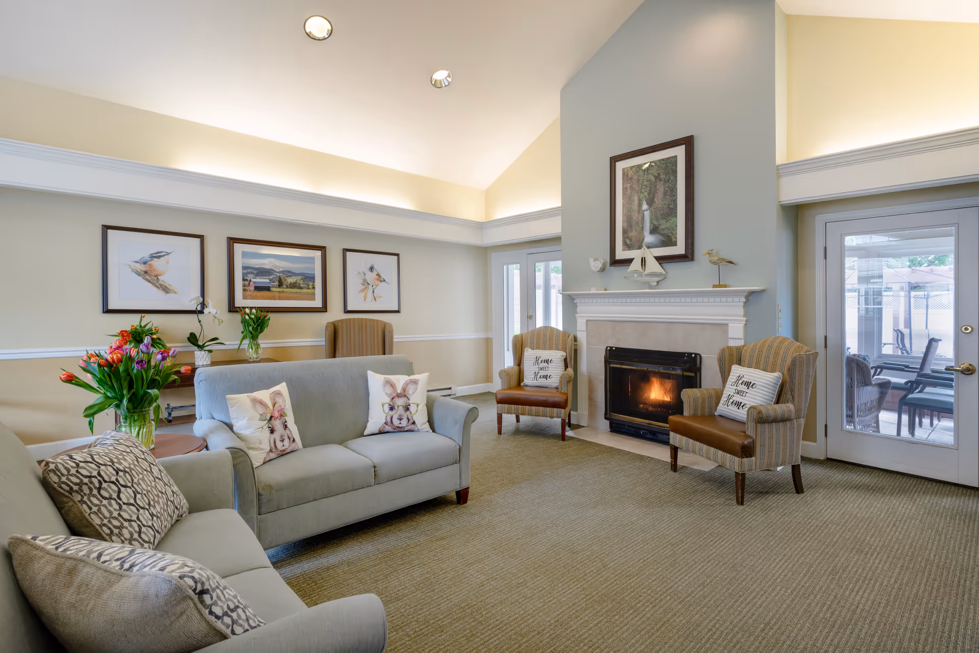 Bright communal living room with sofas, armchairs, a fireplace, framed artwork, and glass doors to the outside.