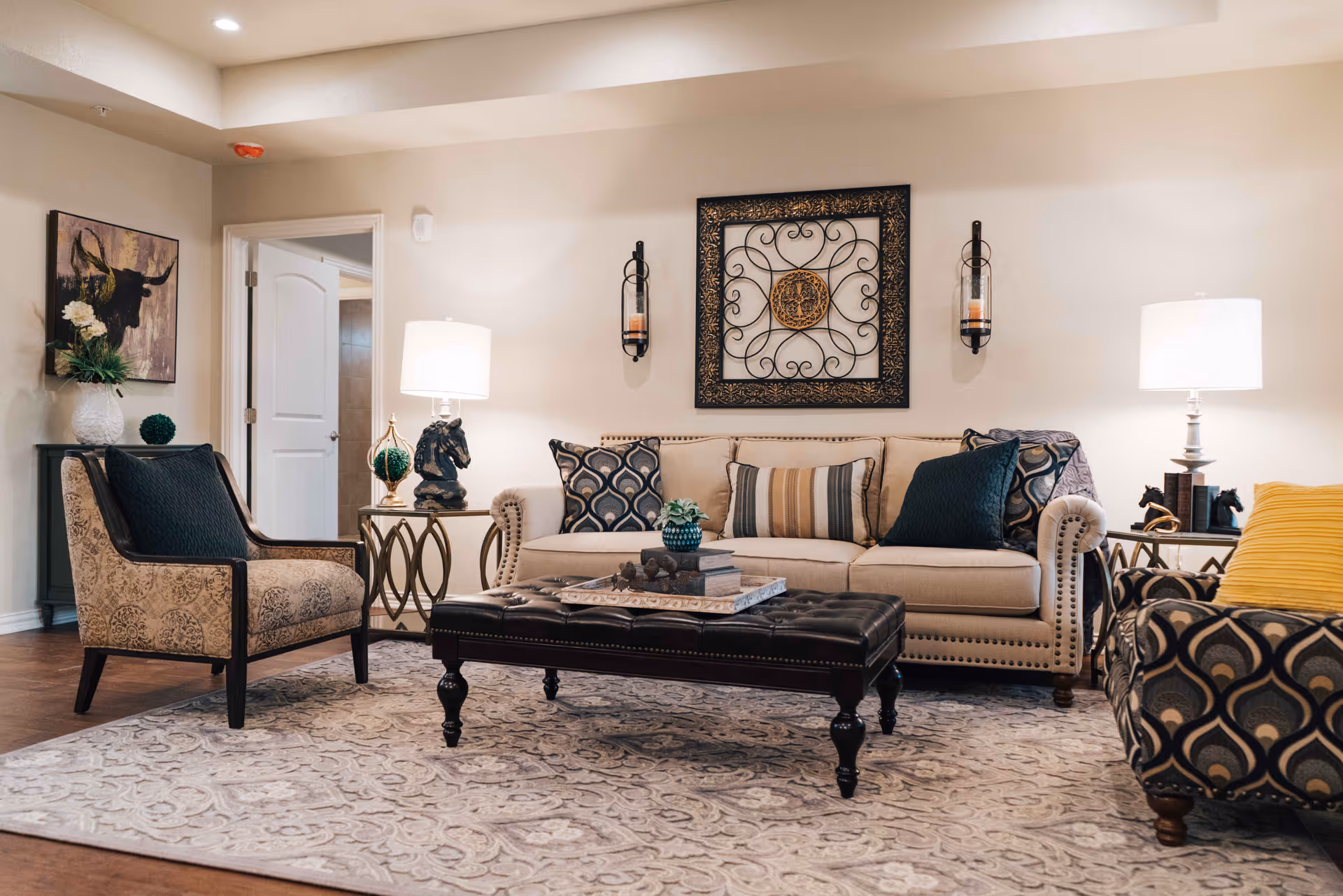 A cozy living room with a beige sofa adorned with patterned and striped cushions, two armchairs with different upholstery patterns, a dark tufted coffee table with books and decorative items, two side tables with lamps and horse head sculptures, a large patterned rug, and wall decorations including a framed metal art piece and a painting of a bull.