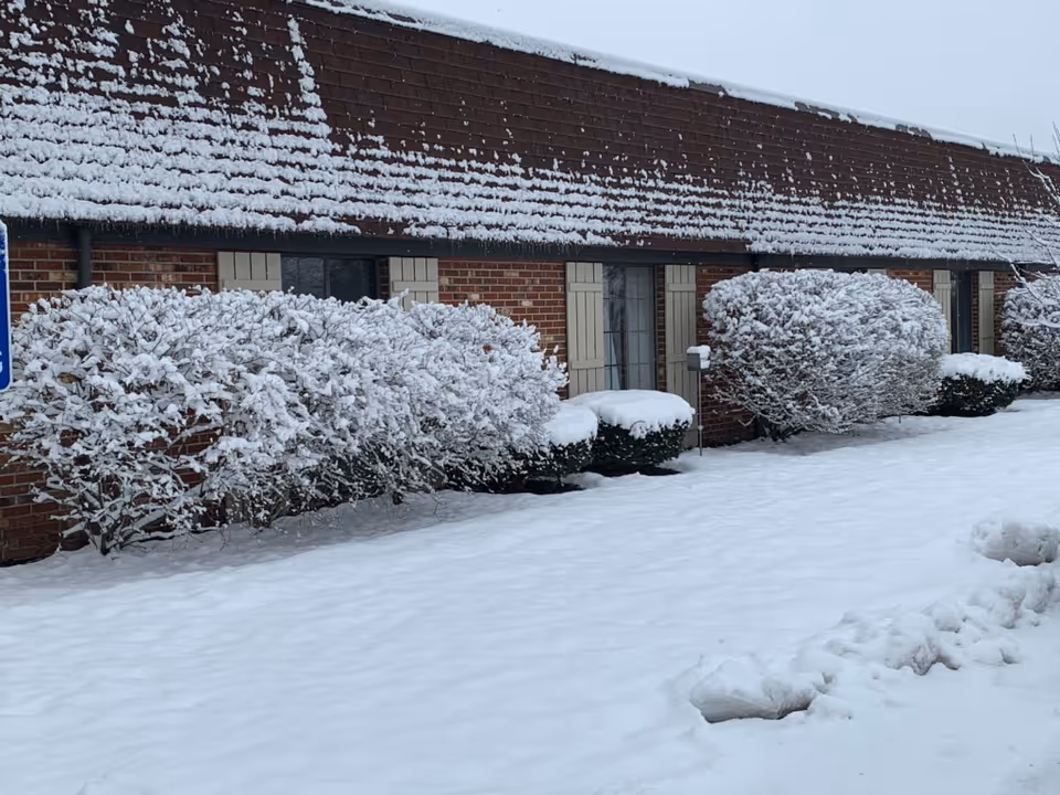 Exterior view of a brick building with a snow-covered roof and snow-covered bushes in front. The ground is blanketed with snow, and the sky is overcast.