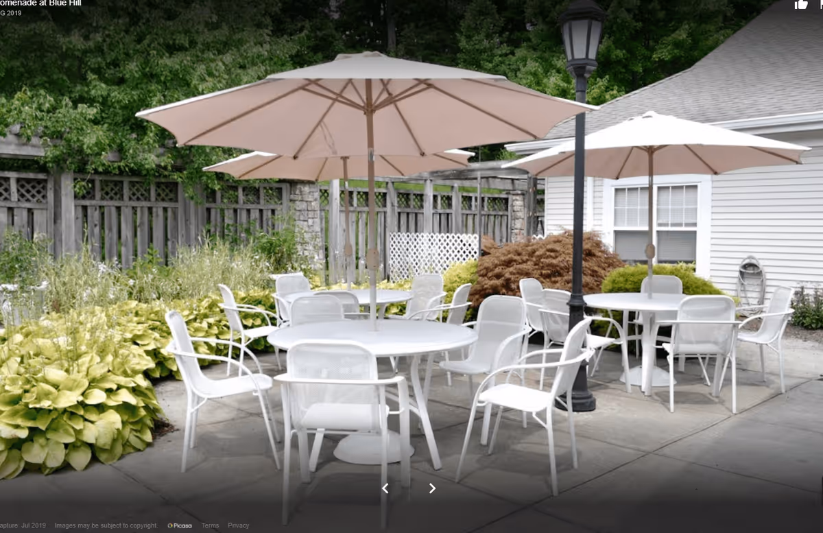 Outdoor patio area with several round white tables and white chairs arranged around them. Each table has a large beige umbrella providing shade. The patio is surrounded by greenery, including bushes and trees, and there is a wooden fence in the background. A lamppost is also visible near one of the tables.