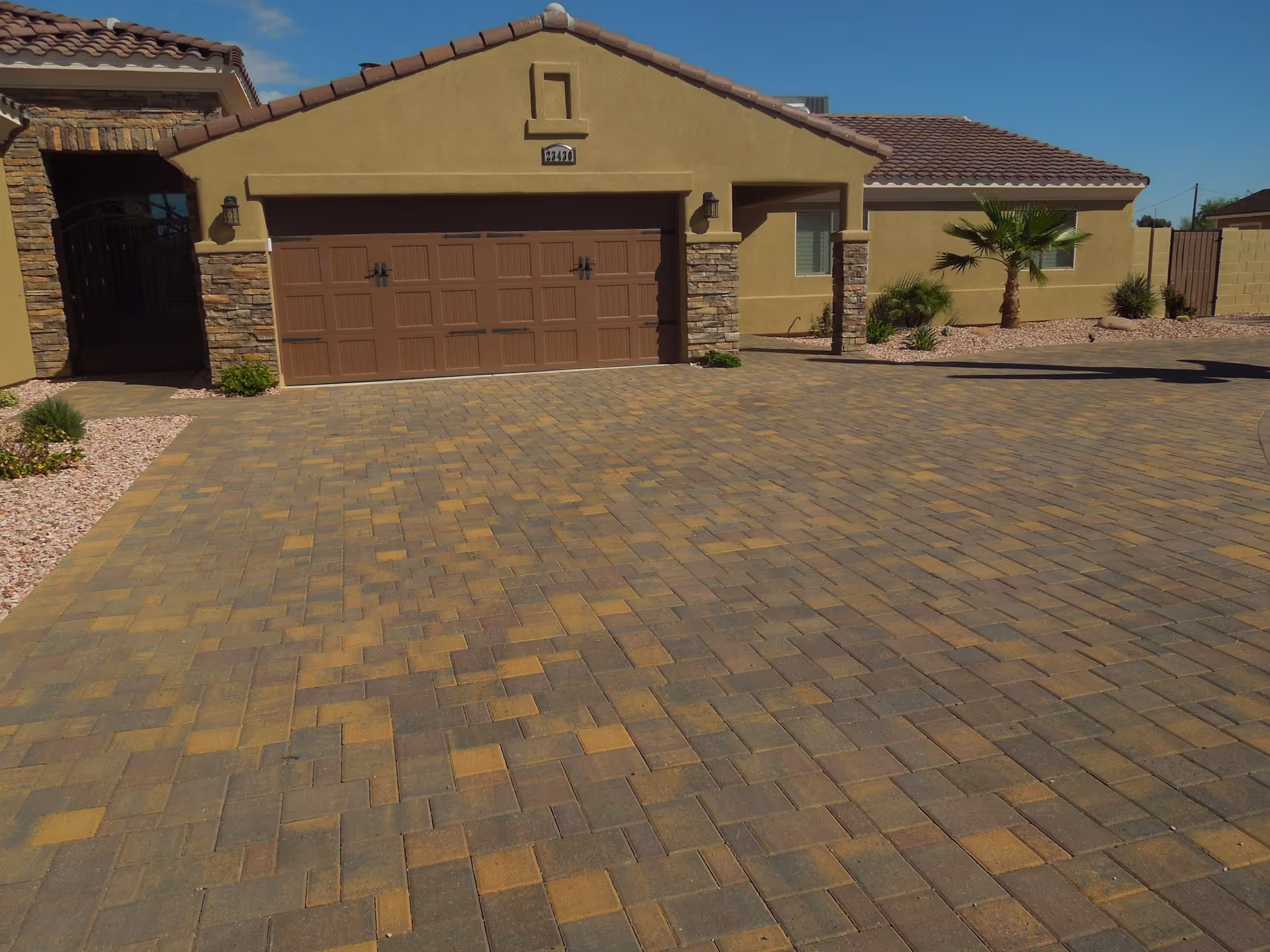 Front exterior view of a single-story house with a brown tiled roof, beige stucco walls, a large brown garage door, and a paved driveway. There are small landscaped areas with rocks, plants, and a palm tree near the house.