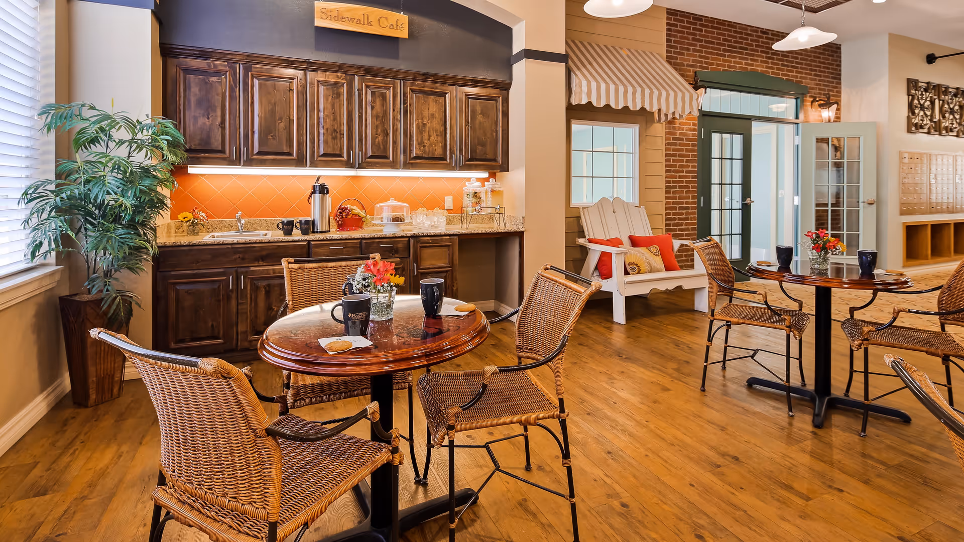 A cozy cafe-style seating area inside a senior living facility with wooden floors, wicker chairs around small round tables, a kitchenette with dark wood cabinets and an orange backsplash, a potted plant near a window with blinds, and a white bench with red and yellow cushions under a striped awning labeled 'Sidewalk Cafe'.