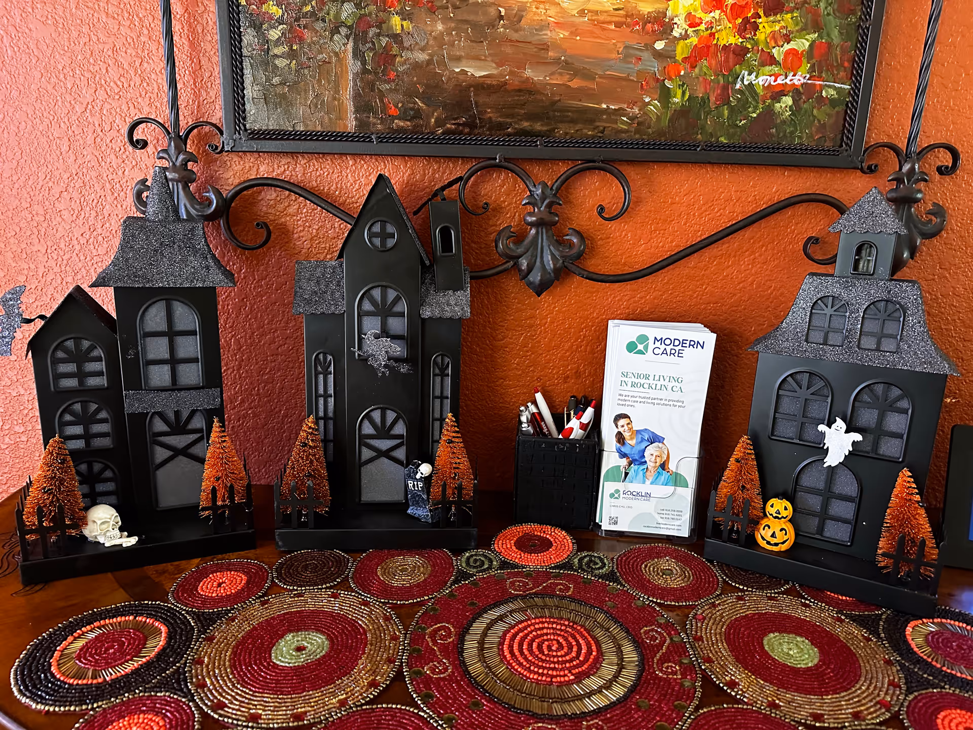 A decorative Halloween-themed display on a table with a colorful circular patterned tablecloth. The display includes three black haunted house models with glittery roofs, small orange trees, a skull, a ghost, pumpkins, and a tombstone. Behind the display is an orange textured wall with a framed painting and a brochure holder containing Modern Care senior living brochures.
