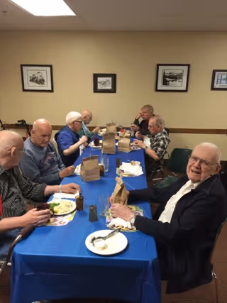 A group of elderly men sitting around a long table covered with a blue tablecloth, eating a meal together in a dining room. There are brown paper bags, plates with food, and drinks on the table. The room has beige walls with framed black and white pictures hanging on them.