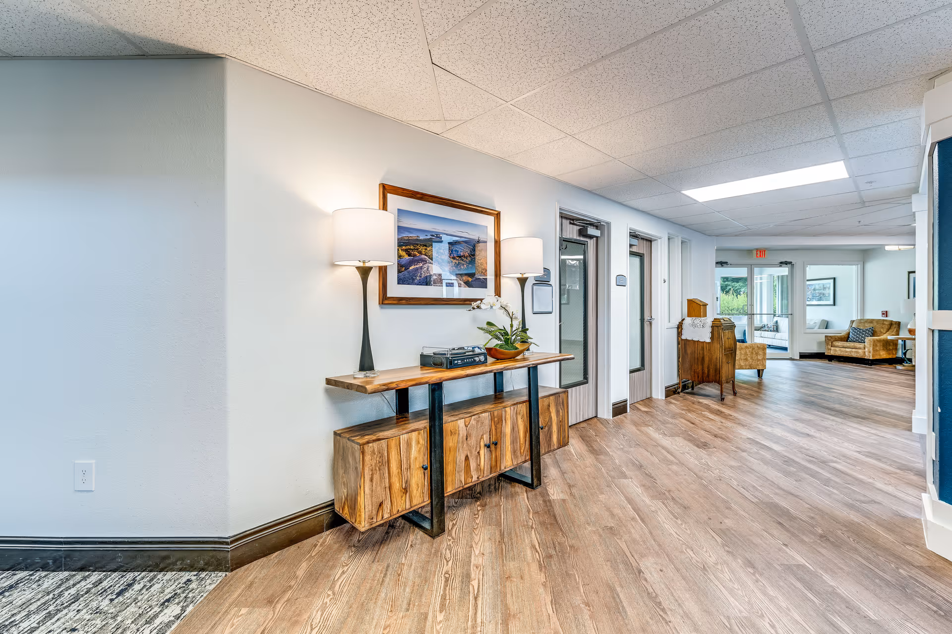 A well-lit hallway in a senior living facility with wood flooring and white walls. There is a wooden console table with two lamps, a decorative plant, and a framed picture hanging above it. The hallway leads to a seating area with armchairs and large windows letting in natural light.