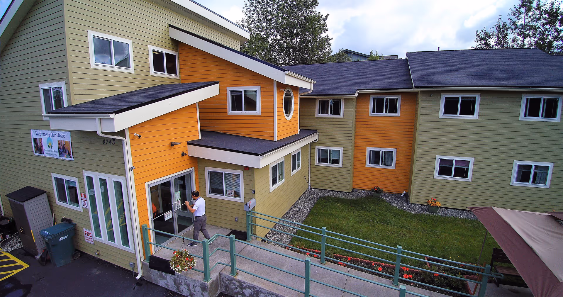 Exterior view of a two-story senior living facility building with green and orange siding, multiple windows, a small lawn with flowers, and a man entering through a glass door. A sign near the entrance reads 'Welcome to Our Home'.