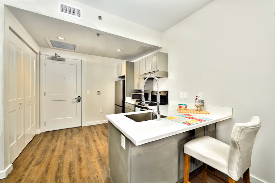 Modern kitchen area with white countertops, a stainless steel sink with a flexible faucet, a stove, microwave, and refrigerator. There is a white cushioned chair at the counter, wooden flooring, and white walls with cabinets above the stove and refrigerator.