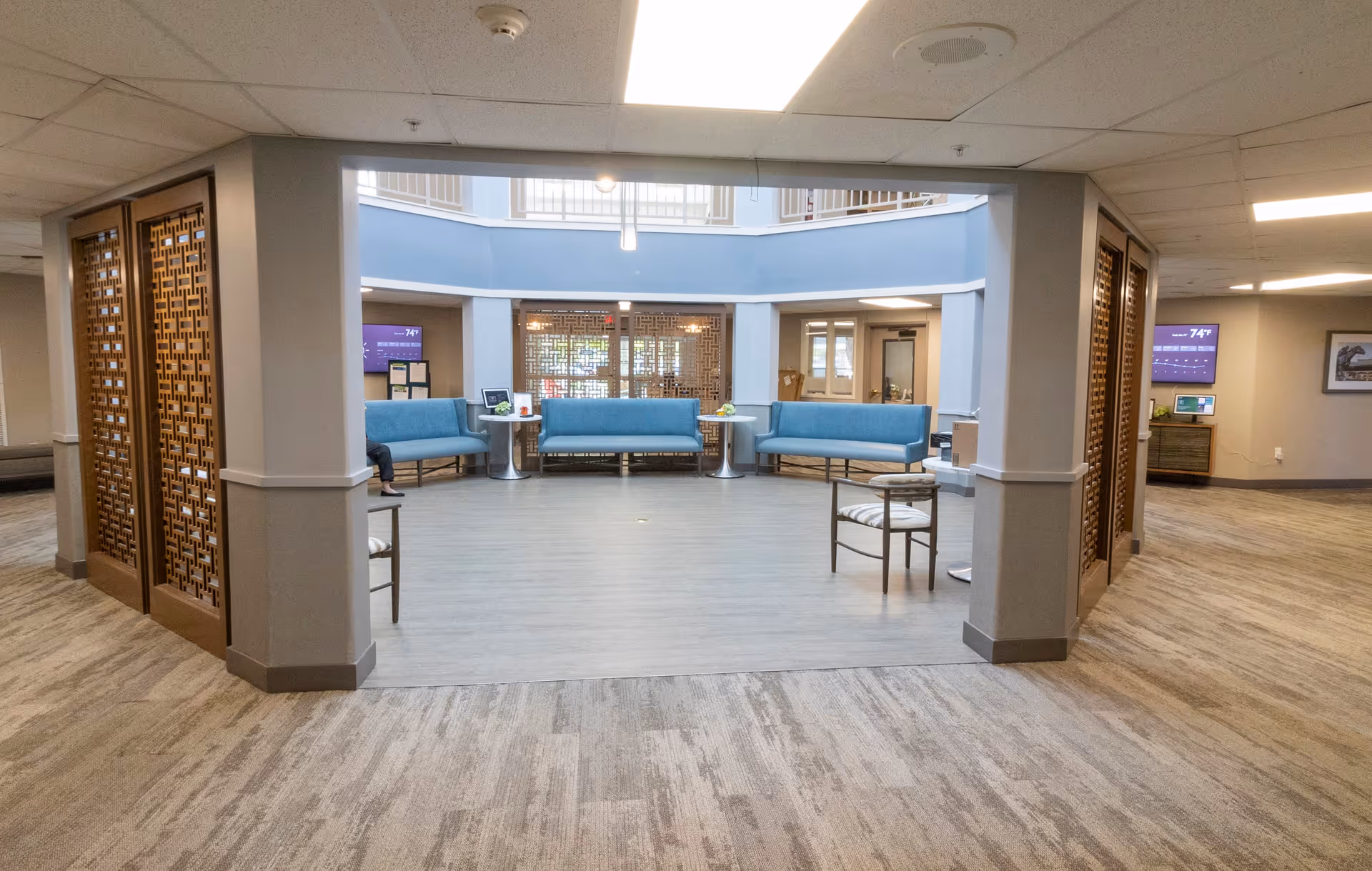 A spacious interior common area with light wood flooring and beige carpet. The area features blue cushioned benches arranged along the walls, two small round tables, and a few chairs with patterned cushions. The space is well-lit with ceiling lights and has decorative wooden lattice panels on the columns. There are digital screens mounted on the walls displaying information.