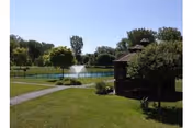 A peaceful outdoor scene at The GreenFields Continuing Care Community featuring a gazebo surrounded by green grass and trees, with a pond and a water fountain in the background under a clear blue sky.