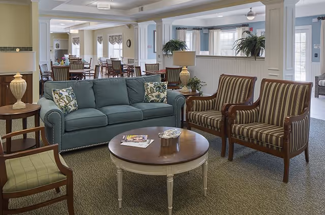 A bright communal living area with a teal sofa, two striped armchairs, a round coffee table, lamps, and dining tables visible in the background.