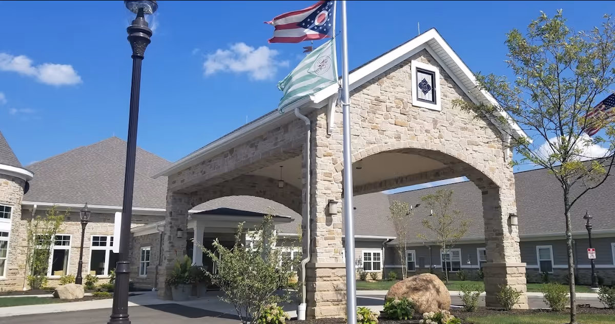 Stone-faced covered entrance and flagpoles at the front of a senior living building with landscaping under a blue sky.