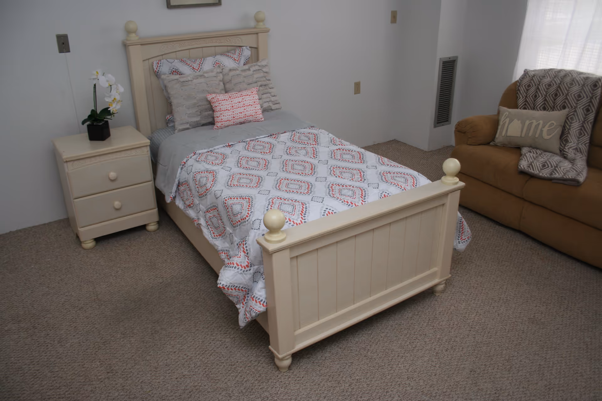 Small bedroom with a single cream bed with patterned bedding, a nightstand with a potted plant, and a sofa against the wall.