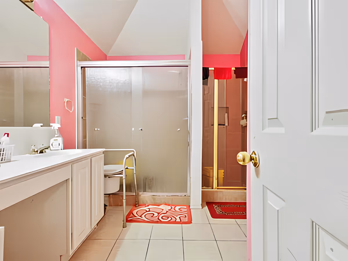 Bathroom with pink walls, a white vanity with a sink and cleaning supplies, a frosted glass shower door, a white commode with a metal support frame, and two red patterned floor mats. The door to the bathroom is partially open with a gold-colored doorknob.