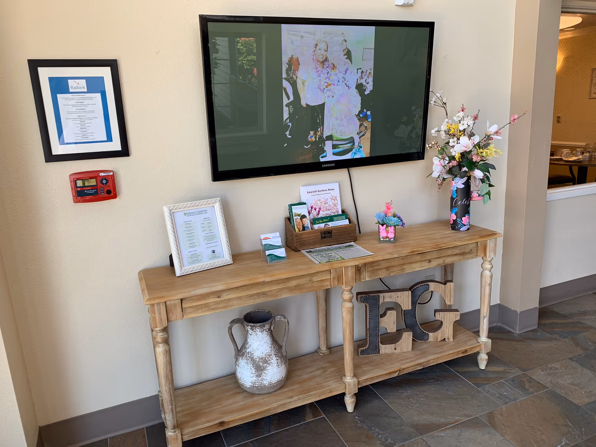 A wooden console table against a beige wall with decorative items including a large ceramic vase, framed documents, brochures, a small flower arrangement, and a tall vase with artificial flowers. Above the table is a mounted flat-screen TV displaying a photo of people. To the left on the wall is a framed certificate and a red fire alarm. The floor is tiled with a stone pattern.