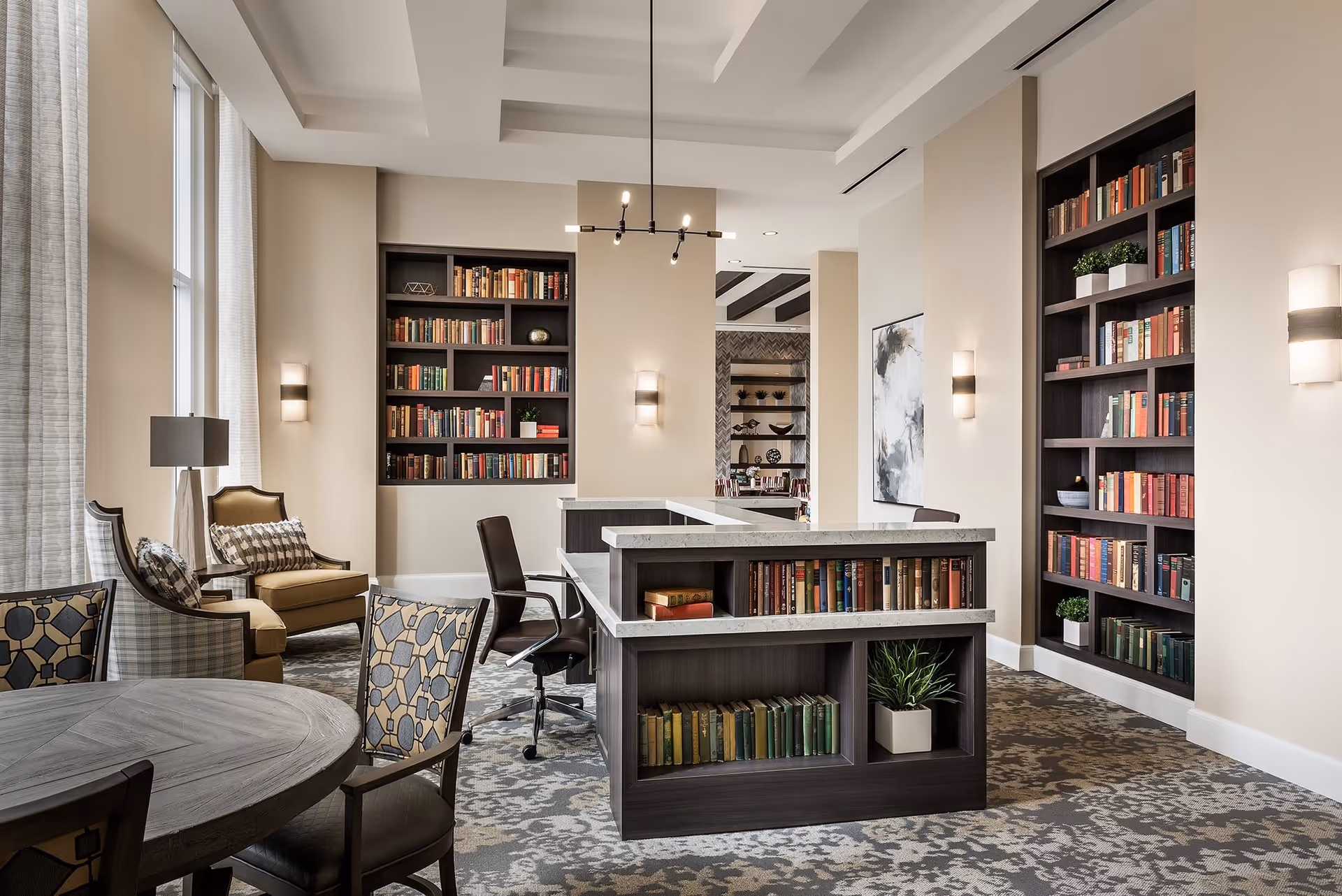 A well-lit library or reading room with built-in bookshelves filled with books and decorative plants. There is a central desk with bookshelves underneath, a round wooden table with patterned chairs, and comfortable armchairs near tall windows with sheer curtains. The room features modern light fixtures and a patterned carpet.