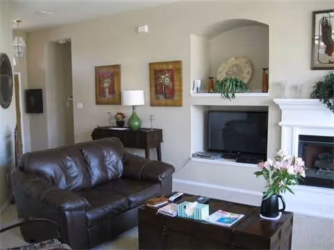 A cozy living room with a dark brown leather loveseat, a wooden coffee table with books and a vase of pink flowers, a flat-screen TV set in a white built-in niche, a fireplace to the right, and a small table with a green lamp and framed artwork on the wall behind the loveseat.
