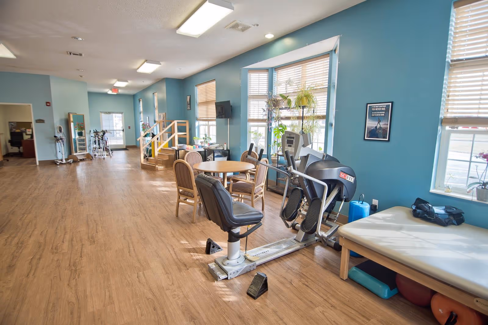 A spacious room with light blue walls and wood flooring featuring exercise equipment including a seated leg press machine and a padded therapy table. There is a round table with four chairs near large windows with blinds, and several plants on the windowsill. The room is well-lit with ceiling lights and natural light from the windows. In the background, there is a small staircase and some medical equipment near an open doorway.