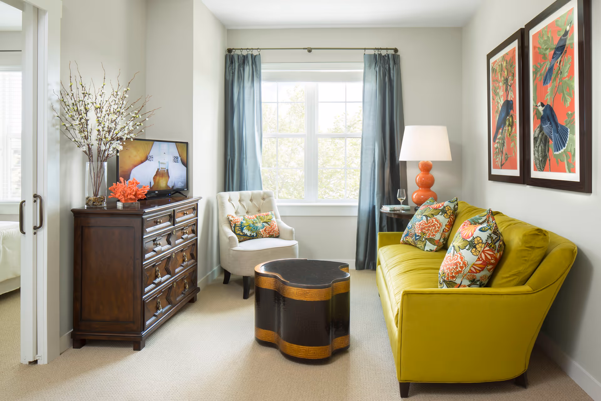 A cozy living room with a bright yellow-green sofa adorned with colorful floral pillows, a unique black and brown coffee table, a beige armchair with a decorative pillow, a wooden dresser with a TV on top, a tall vase with white flowers, blue curtains framing a large window, and two framed bird artworks on the wall.