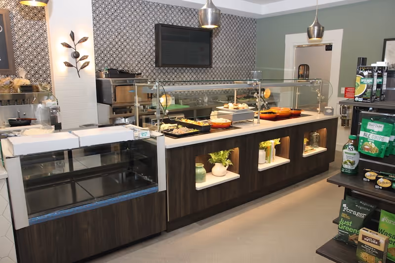 A modern food service counter with glass sneeze guards displaying various dishes in trays and bowls. The counter has built-in shelves with decorative plants and jars. To the right, there is a small shelving unit stocked with packaged snacks and beverages. The background features a patterned wall with a mounted flat-screen TV and pendant lights hanging from the ceiling.