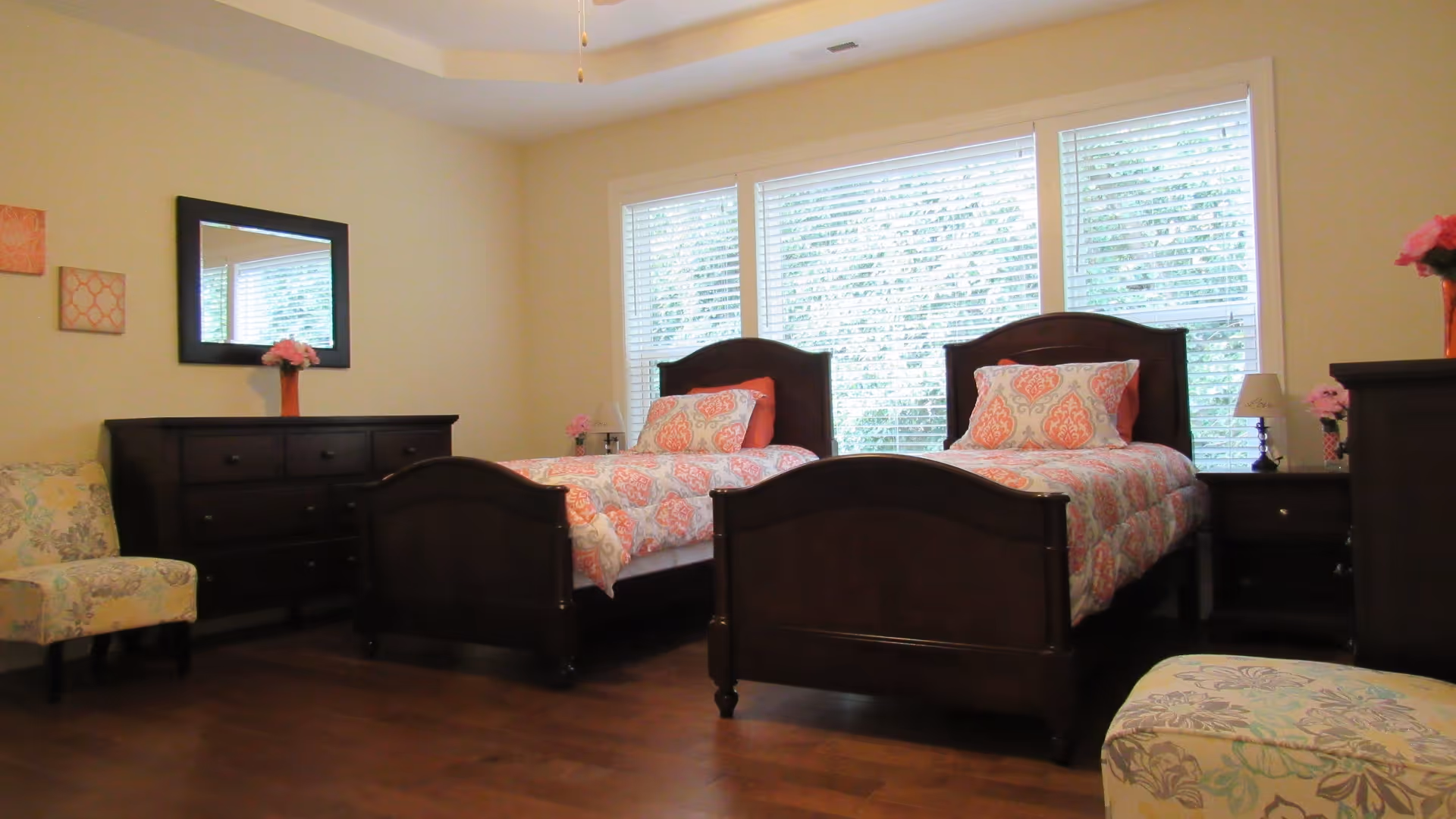 A bedroom with two twin beds featuring dark wooden frames and matching bedding with a coral and white pattern. The room has a large window with white blinds, a dark wooden dresser with a mirror above it, a nightstand with a lamp and flowers, and two upholstered chairs with floral patterns.