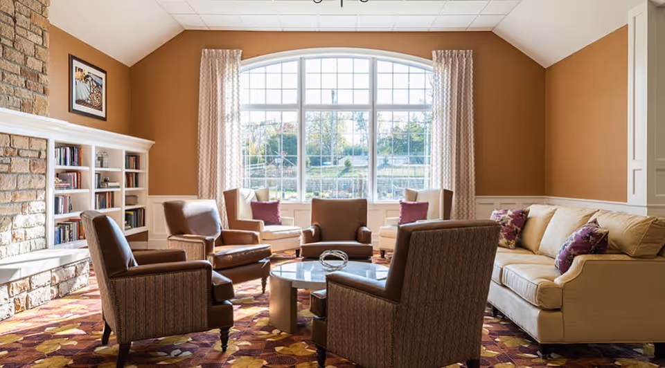Sunlit lounge with armchairs and a sofa arranged around a coffee table in front of a large arched window.