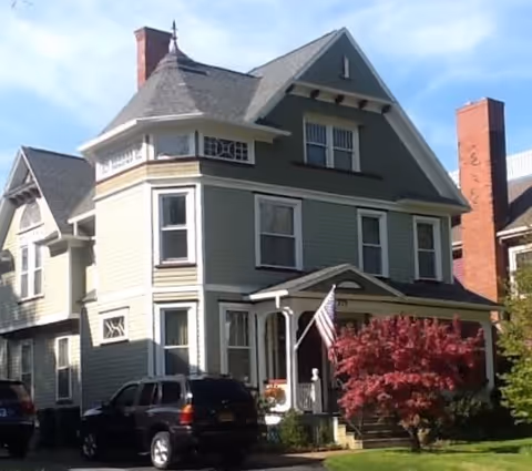 A large, three-story green house with white trim and a steep roof, featuring multiple windows and a small porch with an American flag. There is a red-leafed tree in front and two parked cars on the driveway.
