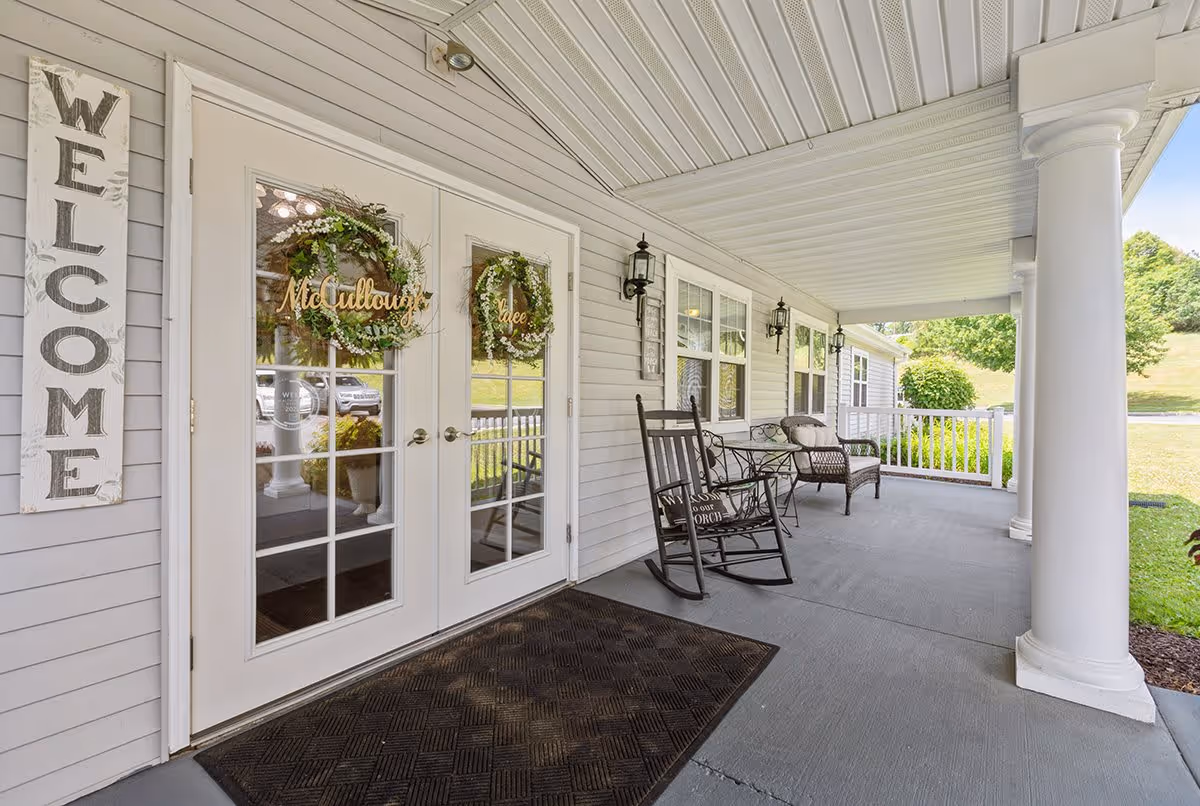 Covered porch area with white columns and a welcome sign on the wall next to double glass doors adorned with wreaths. The porch features a black rocking chair, a small table, and a wicker chair with cushions. Green grass and trees are visible in the background.